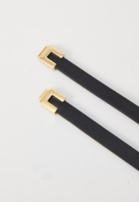 Černé kožené popruhy se zlatými čtvercovými sponami. Textura je hladká a design je minimalistický s moderním vzhledem.