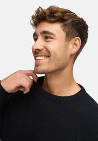 Pull en maille noire avec un col côtelé ; porté par une personne touchant le col, montrant une texture lisse et une coupe ajustée.