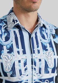 Chemise à manches courtes bleue et blanche à motifs, présentant des designs architecturaux et floraux, avec un devant boutonné et un col structuré.