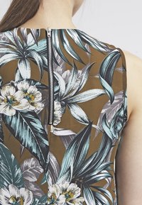 Bezramenný top s květinovým potiskem, který má hnědý podklad, tropické listy v toni modro-zelené a šedé, a stříbrný zip na zadní straně.