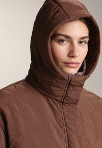 Smeđi kaput s kapuljačom i visokim ovratnikom, od prošivenog materijala s vidljivim šavovima. Ima kontrastnu unutarnju podstavu.