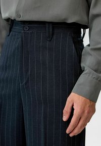Donkere gestreepte broek van textuurstof, met een vlakke voorkant, twee knopen en zijzakken, gecombineerd met een lichtgrijze lange mouw shirt.