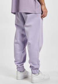 Lichtpaarse trainingsbroek met een relaxte pasvorm, elastische boorden en een gladde textuur, gecombineerd met witte sneakers. Eenvoudig ontwerp, geen zichtbare logo's.