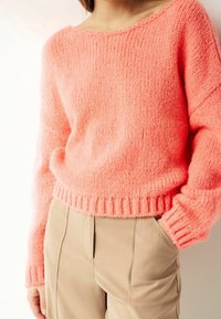 Różowy sweter robiony na drutach z szerokim dekoltem i ściągaczami przy mankietach. Noszony na beżowych spodniach, eksponując miękką teksturę i luźny krój.