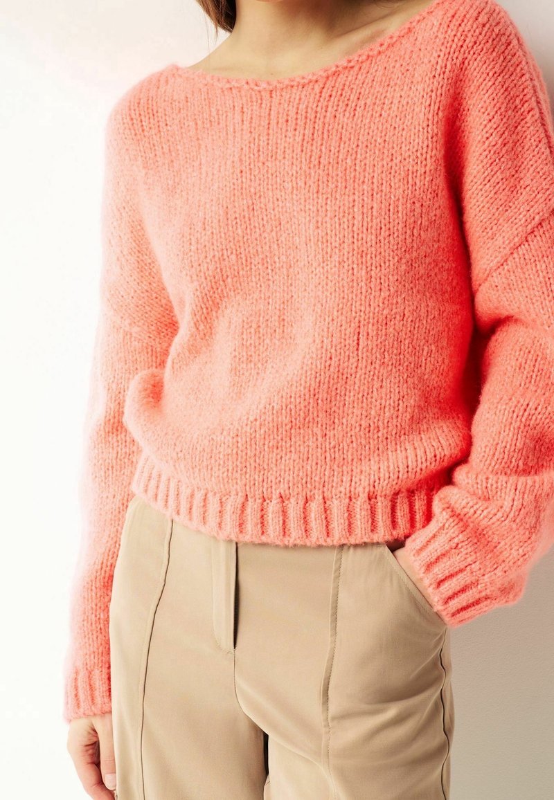 Różowy sweter robiony na drutach z szerokim dekoltem i ściągaczami przy mankietach. Noszony na beżowych spodniach, eksponując miękką teksturę i luźny krój.