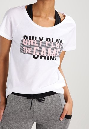 Kobieta w białej koszulce z napisem "ONLY PLAY THE GAME" i szarych spodniach dresowych, pozująca z jedną ręką w kieszeni na neutralnym tle.