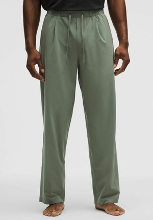 Mand iført olivengrønne løstsiddende bukser med elastik i taljen og snøre, står barfodet mod en ensfarvet baggrund.