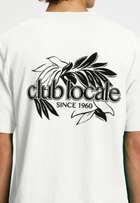 Fehér pamut póló, fekete levél grafikai mintával, a hátsó részén a "club locale" és a "SINCE 1960" felirattal.
