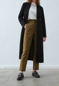 Lange zwarte jas, beige top en olijfgroene broek. Zwarte schoenen met gespdetail. Soepele texturen en een op maat gemaakte pasvorm. Minimalistisch design.