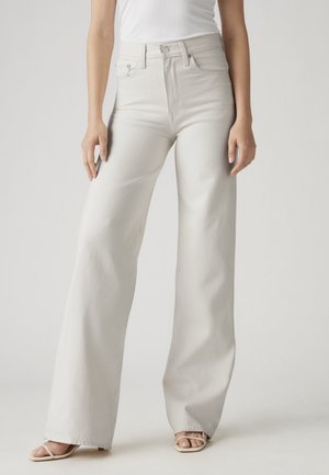 Femme portant un jean blanc taille haute à jambes larges, associé à un haut blanc sans manches et des sandales beiges à bout ouvert.