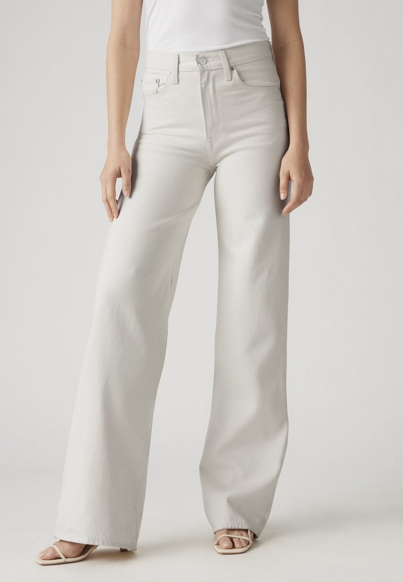 Femme portant un jean blanc taille haute à jambes larges, associé à un haut blanc sans manches et des sandales beiges à bout ouvert.