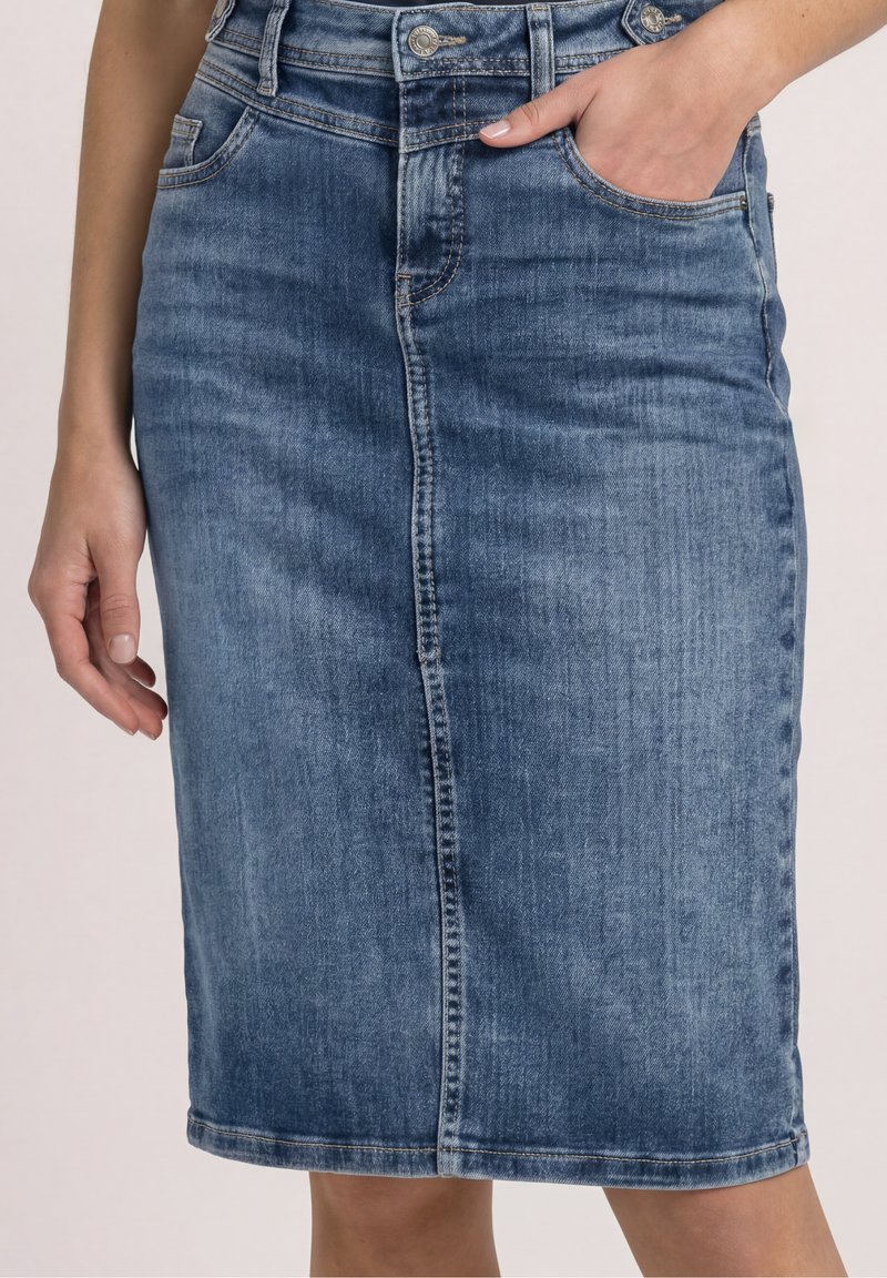 Femme portant une jupe crayon en jean bleu avec couture frontale et passants de ceinture, une main dans la poche et l’autre détendue sur le côté.
