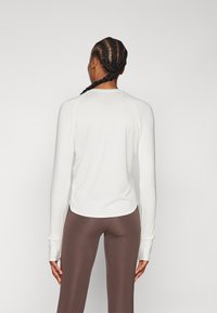 Fehér, hosszú ujjú felső, lekerekített aljjal és raglán ujjal, barna leggings-szel párosítva, sima textúrával és testhez simuló kialakítással.