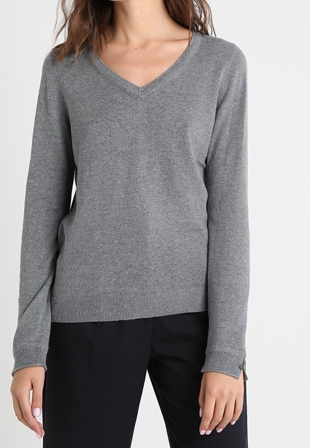 Femme portant un pull gris à col en V à manches longues et un pantalon noir, debout devant un fond clair uni.