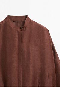 Chemise en lin marron avec un col montant et un seul bouton marron visible au niveau du col, montrant la texture et des détails de couture minimaux.