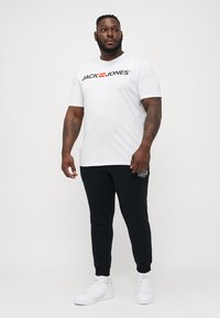 Witte t-shirt met zwart logo "JACK & JONES", gecombineerd met zwarte joggingsbroek met een geborduurde accent. Schoeisel: witte sneakers.