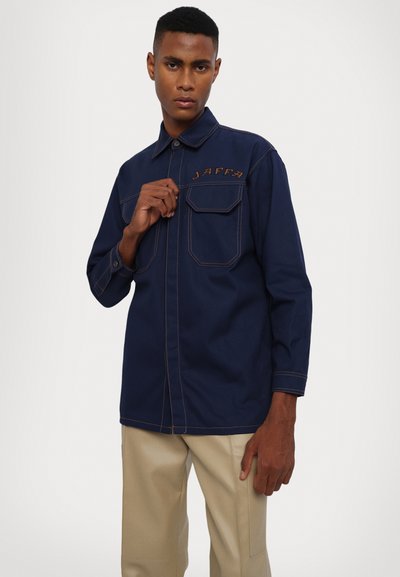 Maglietta a maniche lunghe blu navy con due tasche sul petto, cuciture a contrasto e la parola "JAFFA" ricamata in beige sul lato sinistro.