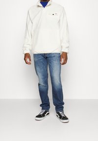 Sudadera de forro polar blanco con cuello alto, bolsillo frontal, combinada con unos vaqueros de mezclilla azules y zapatillas de deporte en blanco y negro.