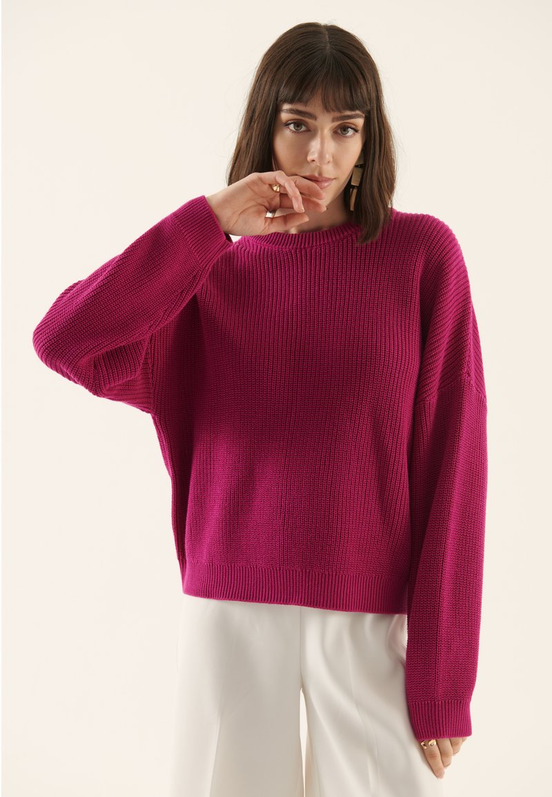Pull en tricot fuchsia avec un col rond, une texture côtelée et des épaules tombantes, associé à un pantalon large de couleur claire.