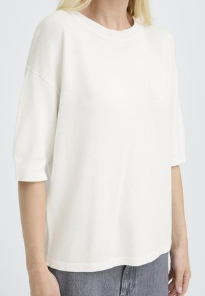 Weißes Strickoberteil mit kurzen Ärmeln, rundem Ausschnitt, lockerem Schnitt und gerippten Bündchen, kombiniert mit dunkelgrauen Jeans. Glatte Textur.