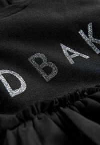 Černá tkanina se shrnutým lemem obsahuje stříbrné glitrové písmo tvořící slovo "DBAK". Hladký povrch s jemným konečným zpracováním zvýrazňuje živé detaily.