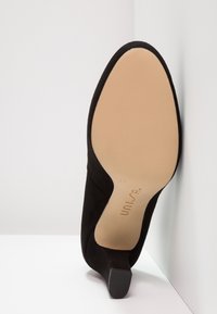 Black suede high heel shoe with a pointed toe and a slim, angled heel. The sole is beige, featuring a textured grip and a size label.