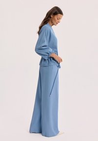 Kobieta ubrana w luźną, niebieską bluzkę z długim rękawem ze ściągaczami na mankietach oraz pasujące szerokie spodnie, stojąca bokiem na jednolitym tle.