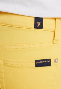 Jeans de mezclilla amarillos con una textura suave, que presentan una etiqueta en la cinturilla de color navy con el número "7" y una etiqueta de marca que dice "para toda la humanidad".