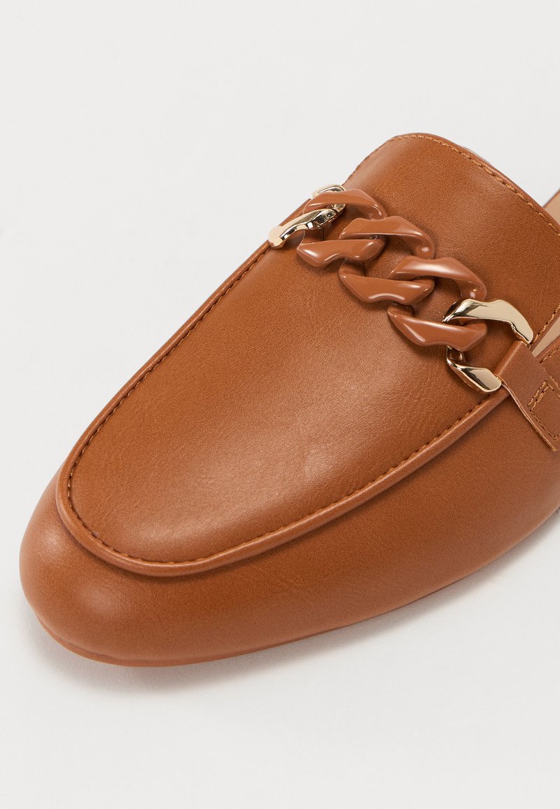 Mocassin en cuir tan à bout rond, présentant un détail de chaîne épaisse avec des accents en métal brun et or. Texture lisse, design minimaliste.