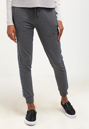 Person trägt dunkelgraue Jogginghosen mit gemusterten Seitenstreifen und schwarzen Slip-on-Schuhen mit weißen Sohlen.