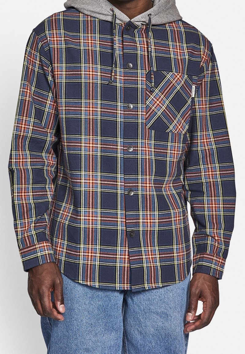 Koszula na guziki w kratę w kolorach granatowym, czerwonym, żółtym i zielonym. Posiada kieszeń na piersi i kaptur z regulowanym sznurkiem. Tkanina wydaje się miękka i lekko fakturowana.