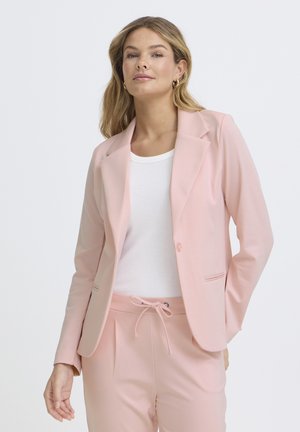 Woman wearing a light pink blazer and matching pants with a white ribbed top, standing against a plain light background.