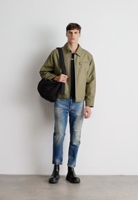 Khaki jakke, svart t-skjorte, slitte blå jeans og svarte gummistøvler. Modellen bærer en svart skulderveske. Minimalistisk design.