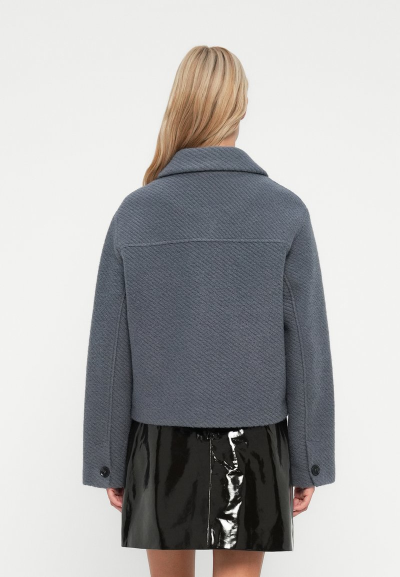 Blonde Frau in einer strukturierten grauen kurzgeschnittenen Jacke und einem glänzenden schwarzen Lackleder-Minirock, die vor einem weißen Hintergrund steht.