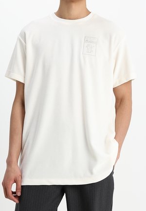 Mand iført en almindelig hvid kortærmet T-shirt med lille tydeligt logo på brystet, kombineret med mørke bukser med fine striber.