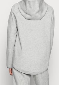 Ljusgrå hoodie med rundad nederkant och en tajt huva. Har en mjuk textur och raglanärmar. Burits med matchande grå byxor.