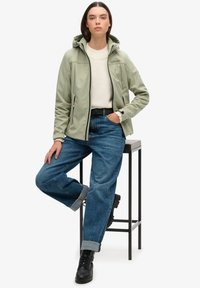 Lichtgroene waterdichte jas met capuchon, ritsluiting en logo, gecombineerd met opgerolde donkerblauwe jeans en zwarte veterschoenen.