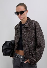 Woman wearing black sunglasses, brown leopard print jacket, black crop top, gray jeans, holding large black leather bag against plain background.