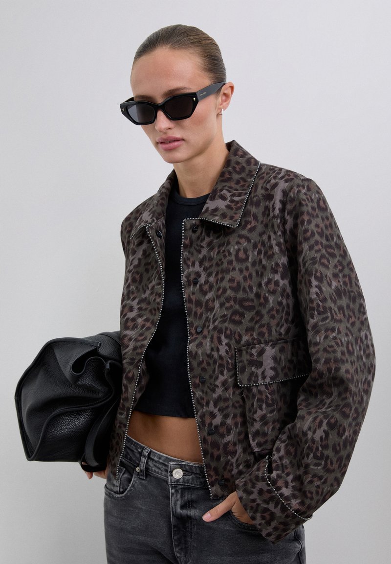 Woman wearing black sunglasses, brown leopard print jacket, black crop top, gray jeans, holding large black leather bag against plain background.