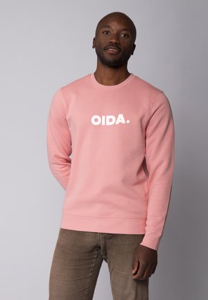 Lichtroze sweatshirt van zachte stof, met een ronde halslijn en witte tekst "OIDA." op de voorkant. Combinatie met bruine corduroy broeken.