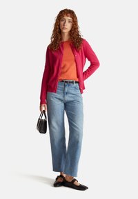 Kobieta z kręconymi rudymi włosami, ubrana w różowy kardigan na pomarańczowej bluzce, jasno-niebieskie dżinsy, czarne baleriny, trzymająca małą czarną torebkę.