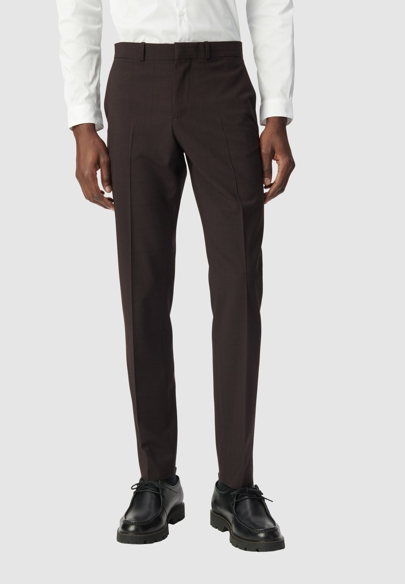 Pantalons marron sur mesure avec un motif de carreaux subtil, fabriqués en tissu doux. Associés à des chaussures en cuir noires et une chemise blanche.