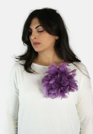 Kobieta o długich ciemnych włosach, ubrana w biały top ozdobiony dużym fioletowym piórem na piersi, patrząca w dół i w bok.