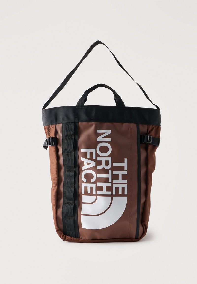 Καφέ και μαύρη τσάντα tote της North Face με μεγάλο λευκό λογότυπο, ρυθμιζόμενο ιμάντα ώμου και δύο κοντές χειρολαβές σε λευκό φόντο.