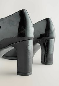 Tocuri de piele lăcuită neagră cu toc bloc. Suprafață netedă, vârf rotund și cusături vizibile pe margine, evidențiind detalii de design minimale.