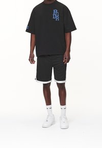 Černé oversized tričko s modrou vyšitou logem, spárované s černými šortkami s bílým lemem. Zahrnuty bílé tenisky a ponožky po kotníky.