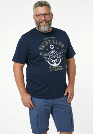 Lächelnder bärtiger Mann mit Brille, trägt ein marineblaues Yachtclub-T-Shirt mit nautischem Design und blaue Cargoshorts, steht vor weißem Hintergrund.