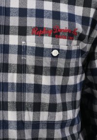 Flanellen shirt met een blauw, zwart en wit ruitpatroon, witte knoopsluiting, linkerkant zak en een rood geborduurd logo.