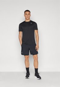 Camisa atlética de manga corta negra y pantalones cortos negros con el logotipo de Under Armour. El modelo, que lleva calcetines negros y zapatillas deportivas, está de pie contra un fondo blanco.