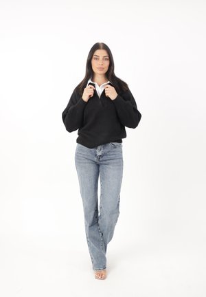Maglione nero a trama con scollatura a V, abbinato a jeans azzurri a vita alta e tacchi trasparenti open-toe. Camicia bianca con colletto visibile.